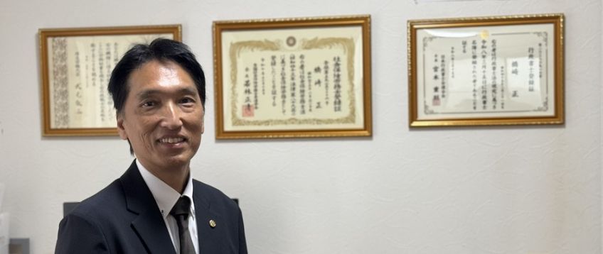 事務所に掲げた社会保険労務士と行政書士の登録証の前に立つ はしざき事務所 代表 橋崎 正